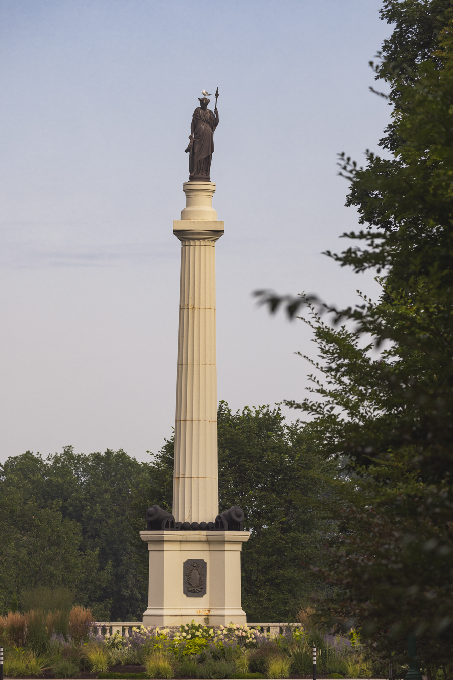 Image of the Monument des Braves located in Parc des Braves, Quebec City, which is managed by the Plains of Abraham. The monument, standing 22 meters high, commemorates the Battle of Sainte-Foy on April 28, 1760. It features a statue of Bellona, the Roman goddess of war, atop a column, with plaques at the base honoring the generals of the opposing armies, Lévis and Murray. The monument is surrounded by lush greenery, highlighting its historical significance and beauty.